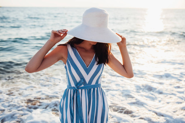 Ελκυστική, όμορφη γυναίκα με λευκό καπέλο στο κεφάλι στο ηλιοβασίλεμα στη θάλασσα  - Φωτογραφία, εικόνα