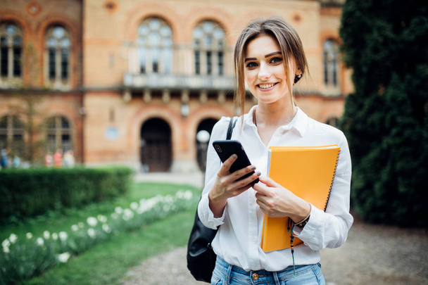 Single student walking and reading mobile phone messages with university building in the background - Фото, изображение