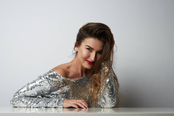 Fashion portrait of a beautiful smiling woman with long hair in a pretty silver dress with tinsel sitting at the table over empty light background. Fashion photo.Horizontal. - Photo, Image
