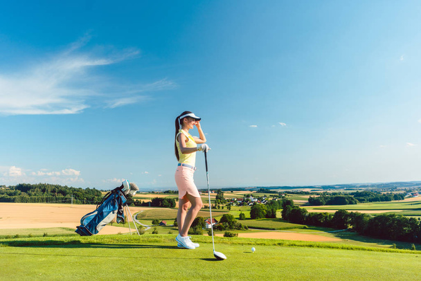Side view of a fit woman wearing modern golf outfits, while looking at the horizon on the green grass of a golf course in the countryside in a sunny day of summer - Photo, Image
