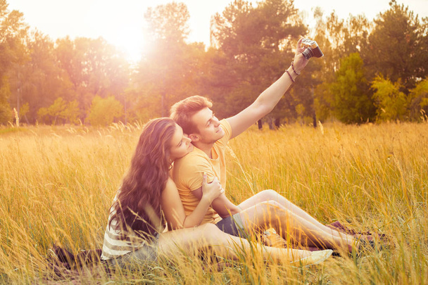 loving couple taking selfie with camera and sitting on floral field in autumnal park at warm sunny day, happiness and love concept - Photo, Image