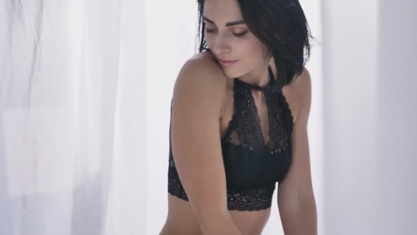 junge sexy kaukasische Brünette in schwarzer Unterwäsche, berührt ihren Körper, schließt die Augen, sitzt auf einer flauschigen Decke, in der Nähe eines Fensters, Tageslicht 50 fps - Filmmaterial, Video
