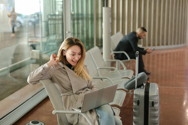 Kaukaski dziewczyny, siedząc w poczekalni z laptopa w pobliżu walizka. - Zdjęcie, obraz