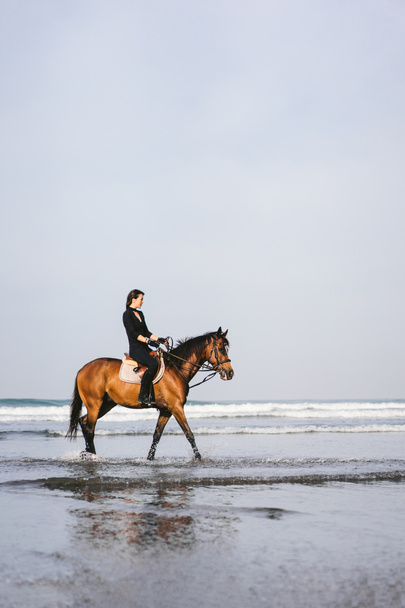 side view of young woman riding horse with wavy ocean behind  - Foto, imagen