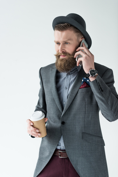 Stylish bearded businessman holding paper cup and talking on phone isolated on light background - Fotó, kép