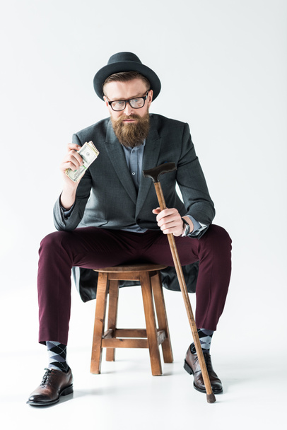 Stylish bearded businessman holding dollars and cane while sitting on stool - Foto, imagen