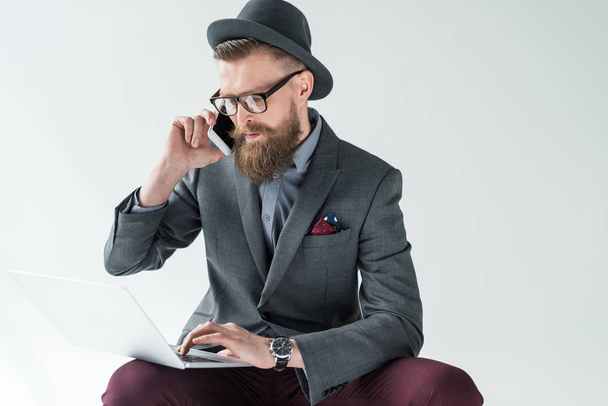 Stylish bearded businessman talking on smartphone and working on laptop isolated on light background - Foto, Imagem