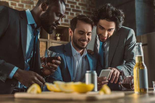 sonrientes hombres multiétnicos usando smartphone y bebiendo whisky juntos
 - Foto, Imagen