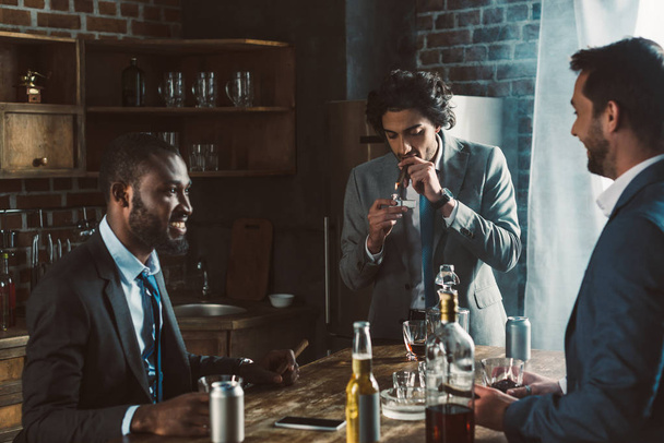 усміхнені молоді друзі-чоловіки в офіційному носінні курять сигари і п'ють алкогольні напої під час вечірки разом
 - Фото, зображення