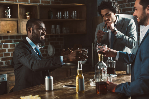 обрезанный снимок улыбающихся многонациональных друзей-мужчин в костюмах, пьющих алкогольные напитки и курящих сигары вместе
 - Фото, изображение