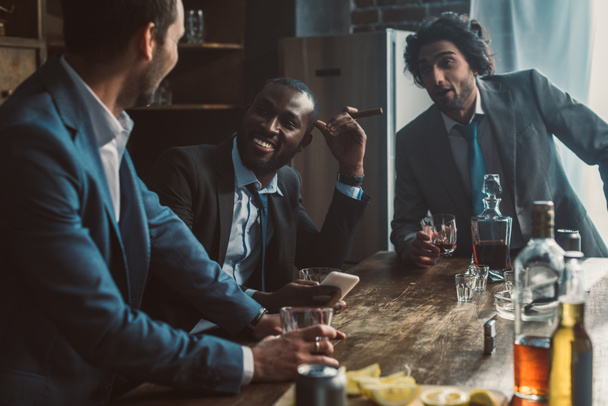 Lächelnde multiethnische Männer, die miteinander reden, Whiskey trinken und Zigarren rauchen - Foto, Bild