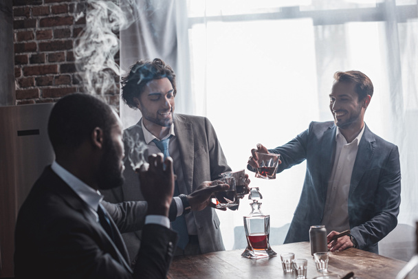 три улыбчивых мультиэтнических бизнесмена курят сигары и звенят стаканы виски
 - Фото, изображение