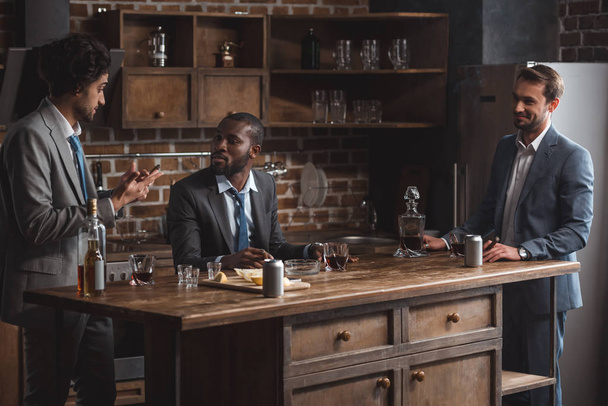 три багатоетнічні друзі чоловічої статі в костюмах розмовляють, п'ючи алкоголь разом
 - Фото, зображення