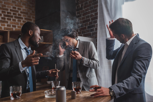 emotionele jonge zakenman sigaar roken en praten door smartphone terwijl vrienden drinken van whisky - Foto, afbeelding