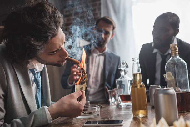 young businessman holding dollar banknote and smoking cigar while partying with friends - Photo, Image