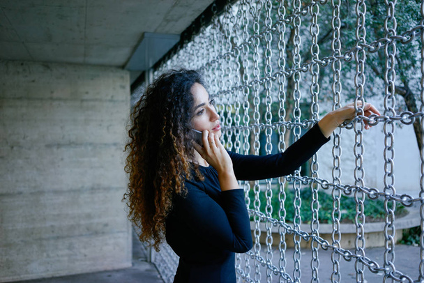young beautiful yooung woman in a black dress with long curly hair talking on phone outdoors - Foto, imagen