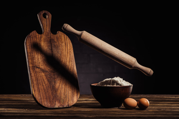flying rolling pin, wooden cutting board, two eggs and bowl with flour on table  - Фото, изображение
