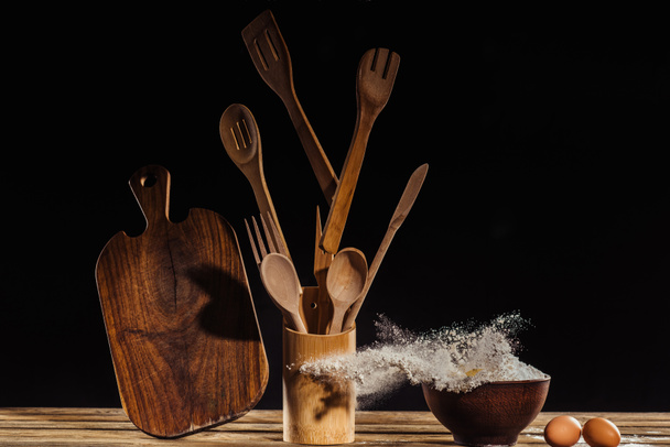 flying cutting board, kitchen utensils, flour from bowl and two eggs on table on black background  - Photo, Image