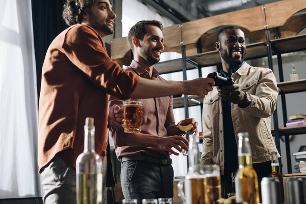 vista de ángulo bajo de los hombres sonrientes multiétnicos jugando con el joystick y beber bebidas alcohólicas juntos
 - Foto, Imagen