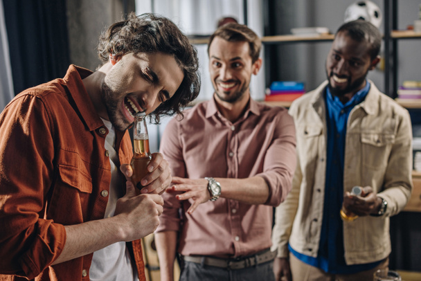 smiling multiethnic men looking at friend opening beer bottle with teeth  - Foto, Bild