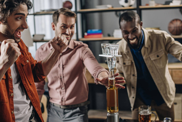 захоплені багатоетнічні чоловіки будують вежу з пляшок і окулярів, розважаючись разом на вечірці
 - Фото, зображення