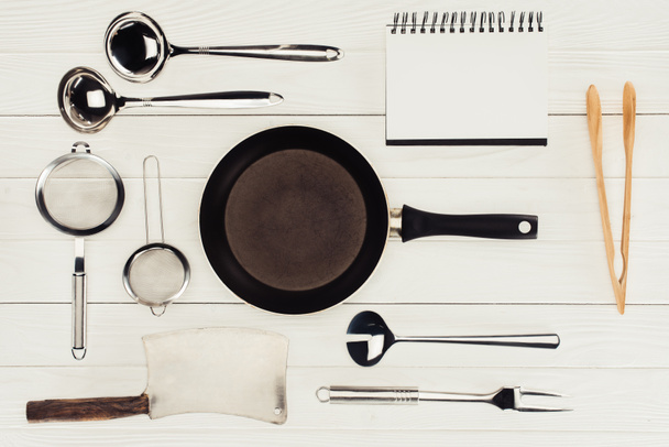 κάτοψη του κενό βιβλίο, τηγάνι, κρεοπώλη τσεκούρι και σκεύη κουζίνας σε λευκό ξύλινο τραπέζι   - Φωτογραφία, εικόνα