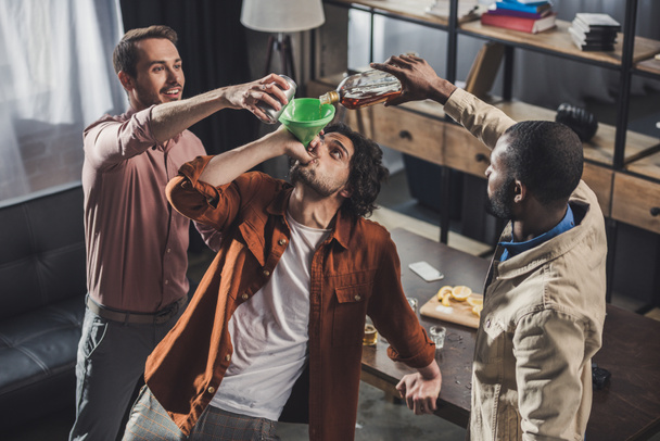 aus der Vogelperspektive: Mann trinkt aus Trichter, während Freunde Alkohol aus Glasflasche und Dose gießen  - Foto, Bild