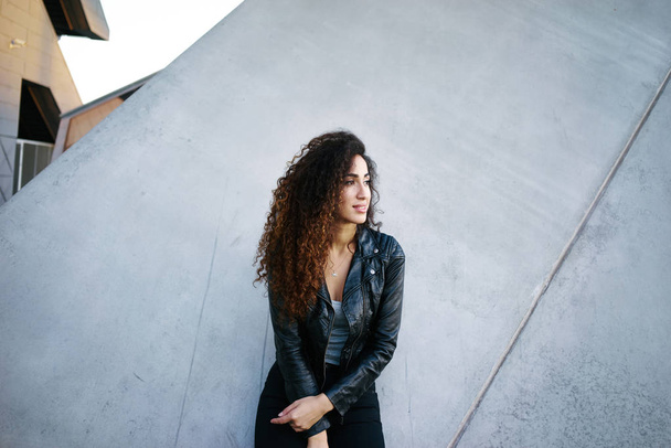 portrait of attractive young woman with long curly hair wearing a black leather jacket posing against gray concrete wall background on city street - 写真・画像