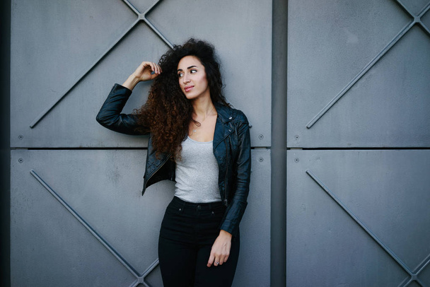 retrato de atraente jovem com cabelos longos encaracolados vestindo uma jaqueta de couro preto posando contra fundo de parede de concreto cinza na rua da cidade
 - Foto, Imagem