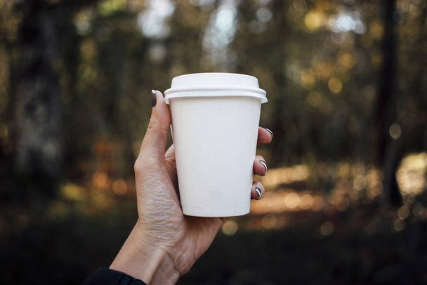 female hand holding a blank paper cup with plastic cap on forest background - Foto, Bild
