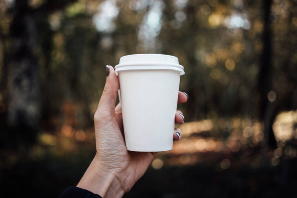 female hand holding a blank paper cup with plastic cap on forest background - Fotoğraf, Görsel
