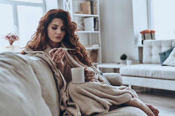 Sick young woman looking at thermometer while sitting on sofa at home               - Foto, afbeelding