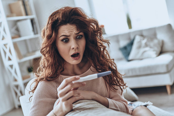 Terrified young woman looking at pregnancy test and keeping mouth open while sitting on sofa at home         - Foto, immagini