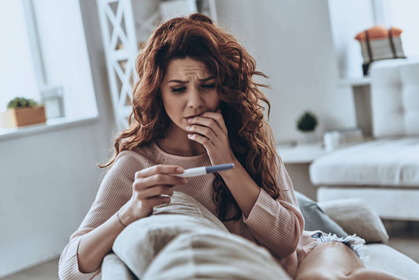 Terrified young woman looking at pregnancy test while sitting on sofa at home           - Foto, immagini