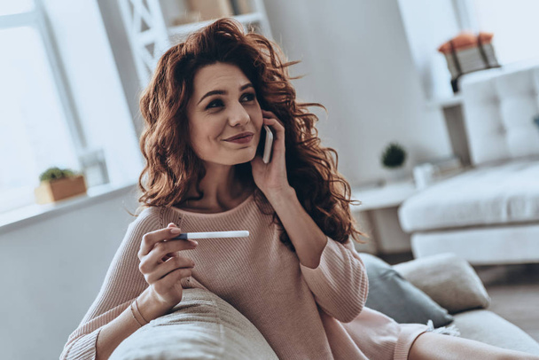 Attractive woman holding pregnancy test and talking on smart phone while sitting on sofa at home         - Photo, Image
