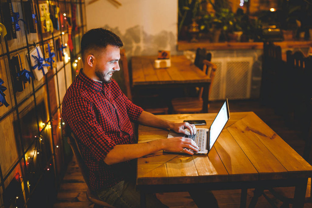 Um jovem caucasiano bonito com barba e sorriso de dente em uma camisa quadriculada vermelha está trabalhando atrás de um laptop cinza sentado em uma mesa de madeira. Mãos no teclado. À noite, no café
 - Foto, Imagem