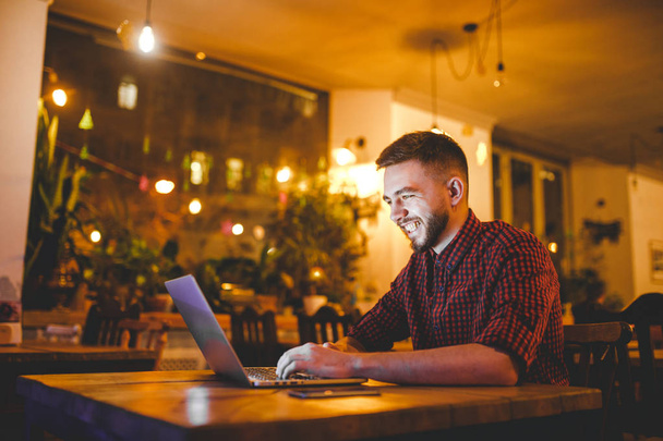A young handsome Caucasian man with beard and toothy smile in a red checkered shirt is working behind a gray laptop sitting at a wooden table. Hands on the keyboard. In the evening at the coffee shop - 写真・画像