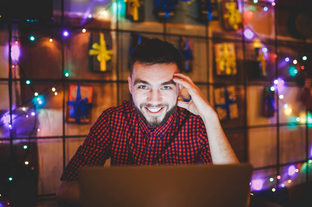 Un joven guapo hombre caucásico con barba y sonrisa dentada en una camisa roja a cuadros está trabajando detrás de un portátil gris sentado en una mesa de madera. Las manos en el teclado. Por la noche en la cafetería
 - Foto, imagen
