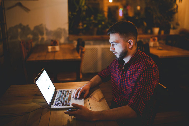 Un joven guapo hombre caucásico con barba y sonrisa dentada en camisa roja a cuadros está trabajando detrás de la computadora portátil sentada en la mesa de madera. Utiliza mantiene el teléfono móvil en la mano. Por la noche en la cafetería
 - Foto, Imagen