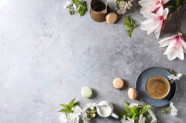Blue cup of black espresso coffee french dessert macaroons, cream and spring flowers magnolia, blooming cherry branches over grey texture background. Top view, space. Spring greeting card, wallpapers - Foto, Imagem