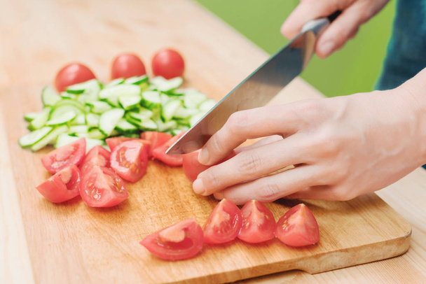 Close-up Delicate female hands cut a large knife with tomatoes on a quarter on a wooden board at home. Home kitchen. Healthy eating. The concept of vegetarianism - Photo, Image