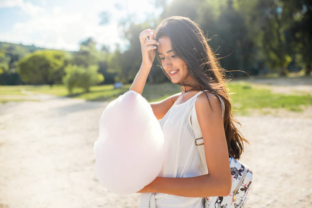 Beautiful portrait of brunette woman with cotton candy and windy hair, with backpack walking in the park and looking at the cotton candy. - Foto, Imagem