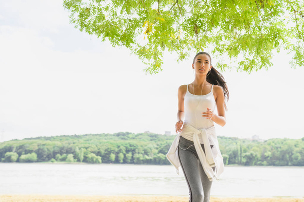 Beautiful brunette female runner running in city park with earphones. Healthy fitness woman jogging outdoors. Nature background. Motivation sport. - Fotoğraf, Görsel