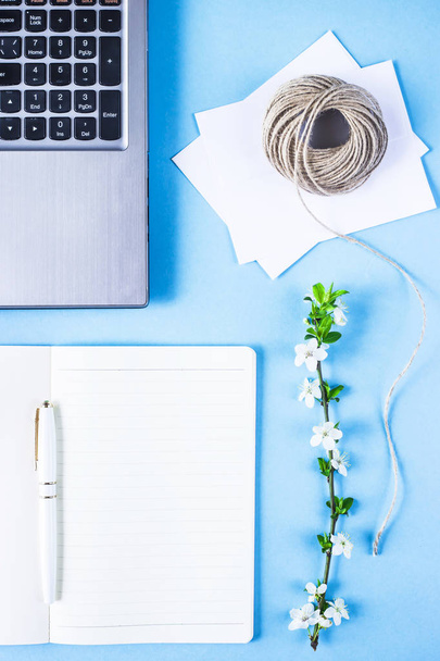kreativer Arbeitsplatz mit Laptop, Notizbuch, Briefumschlägen, Zwirn und blühendem Zweig auf blauem Hintergrund. Frühlingskonzept - Foto, Bild