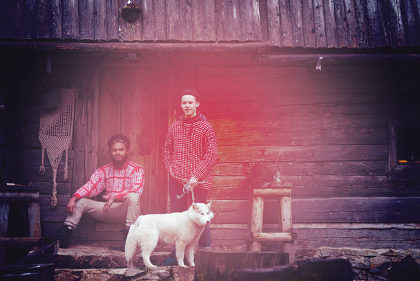 hipsters couple portrait, two young  man with white husky dog  sitting in front of old wooden retro house - Фото, изображение