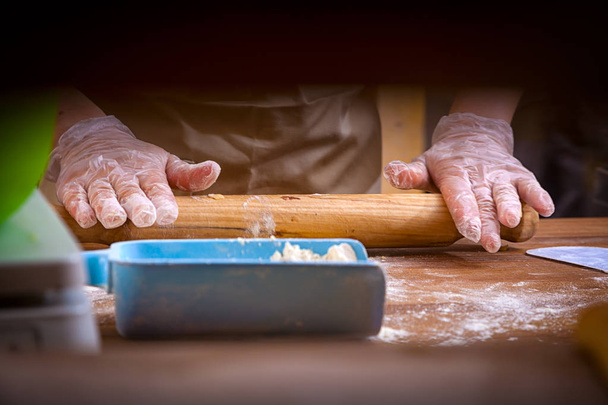A close-up of a male baker in protective gloves pulls out the dough on a knotted table with a rolling pin - Photo, Image