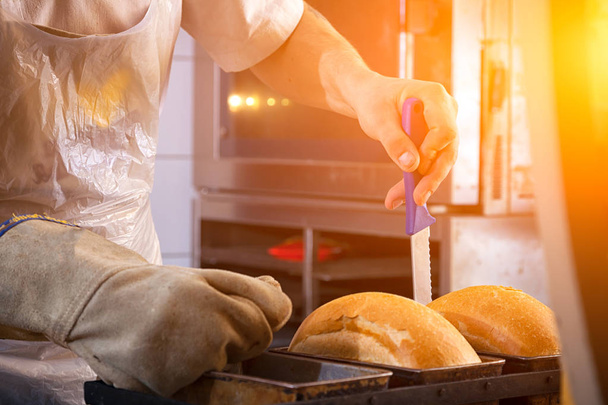 Bliska mężczyzna baker noszenia rękawic ochronnych dotyka świeżo gotowane chleba nożem, który właśnie wziął z pieca w piekarni - Zdjęcie, obraz