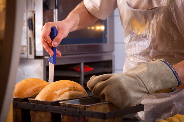 A close-up of a male baker wearing protective gloves touches a freshly cooked bread with a knife, which he just took out of the oven in the bakery - Photo, Image