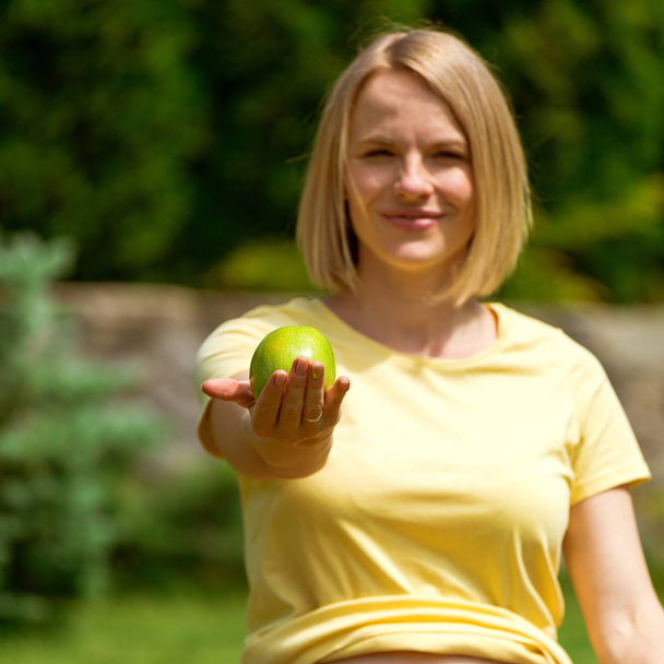 Pregnant woman sitting on grass and holding out apple in hand - Photo, Image