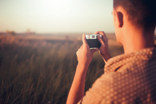 Handsome back view of a young man with vintage camera, on sunset and field background. Autumn mood and summer mood. Photography - Photo, Image